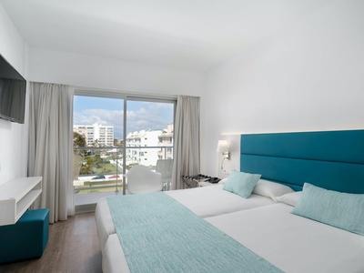 Světly hotelový pokoj s manželskou postelí, tyrkysovými doplňky a balkonem s výhledem na město.