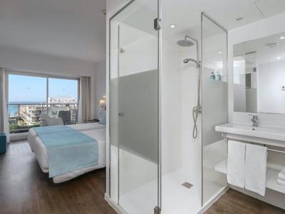 Moderní hotelový pokoj s postelí, velkým skleněným sprchovým koutem a výhledem na město oknem.