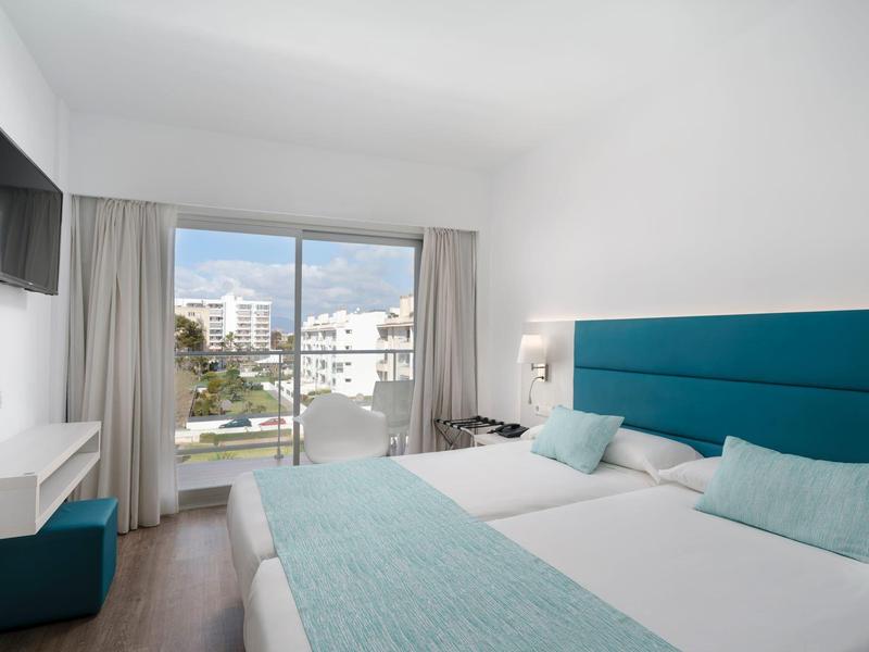 Camera d'albergo moderna con due letti singoli, accenti turchesi e balcone con vista sulla città.
