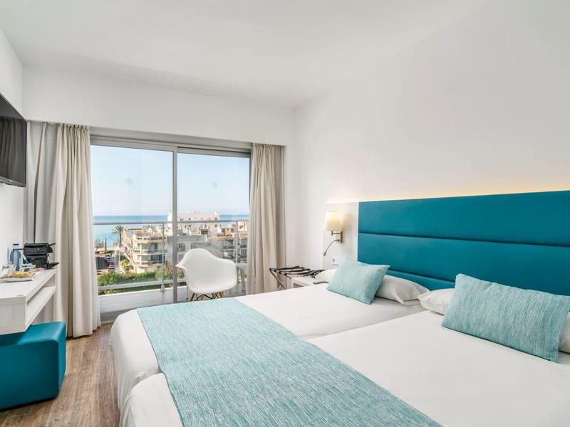 Camera d'hotel moderna con due letti singoli, testiera blu e vista mare attraverso una grande finestra.