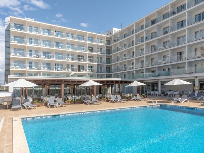 Hotel con grande piscina all'aperto, lettini e ombrelloni sotto un cielo azzurro.