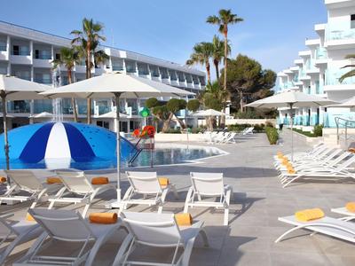 Hotelpoolbereich mit Liegestühlen, Sonnenschirmen und Palmen zwischen weißen Gebäuden.