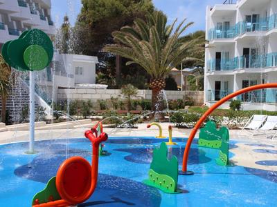 Bunter Kinderspielplatz mit Wasserspielen neben einem Pool und weißen Gebäuden im Hintergrund.