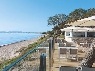 Strand mit ruhigem Meer und Terrasse mit Tischen, Stühlen und Sonnenschirmen unter blauem Himmel.