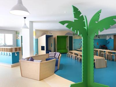 Moderner Kinderraum mit blauen und beigen Sitzmöbeln, Holztischen und großer, grüner Palmen-Deko.