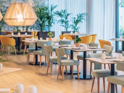 Modernes Restaurant mit hellen Stühlen, Holztischen und großen Pflanzen an Fenstern und Wänden.