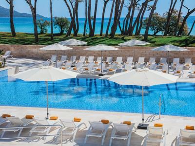 Großer blauer Pool mit weißen Liegestühlen und Sonnenschirmen, im Hintergrund Bäume und Meer.