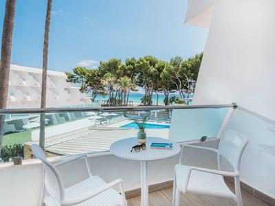 Balkon mit weißem Tisch und Stühlen, Glasgeländer, Blick auf Pool, Bäume und blauen Himmel.