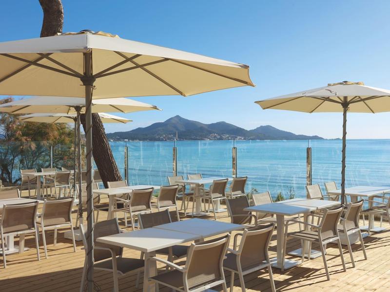 Freiluftrestaurant mit weißen Tischen und Stühlen, großen Sonnenschirmen, Blick auf blaues Meer und Berge.