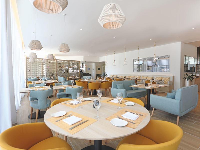 Helles Restaurant mit runden Holztischen, gelben und blauen Sesseln und modernen Lampen.