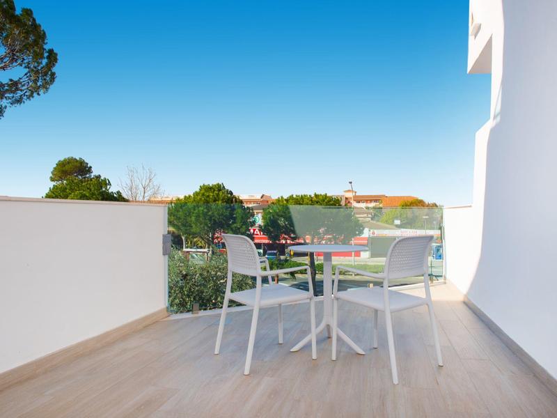 Balkon mit weißem Geländer, Tisch und zwei Stühlen, Blick auf grüne Bäume und blauen Himmel.