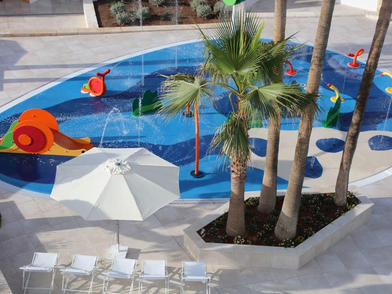 Spielplatz mit blauem Boden, roten Schilden, Palmen und weißen Sonnenschirmen in einem Innenhof.