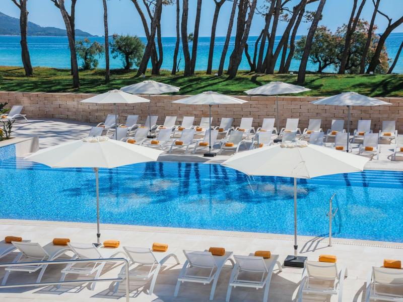 Großer blauer Pool mit weißen Liegestühlen und Sonnenschirmen, im Hintergrund Bäume und Meer.