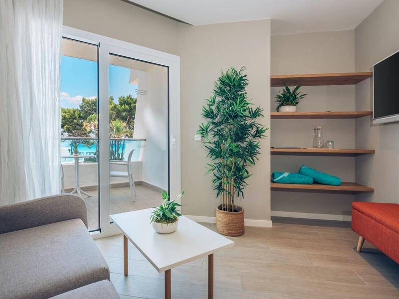 Helles Wohnzimmer mit Sofa, Couchtisch, großem Pflanzenkübel und Balkon mit Poolblick.