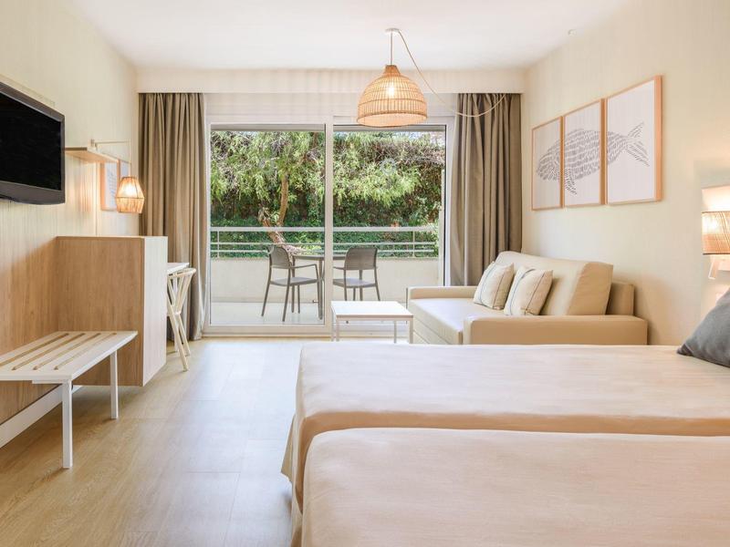 Camera d'albergo luminosa con due letti, divano e terrazza con vista sul verde.