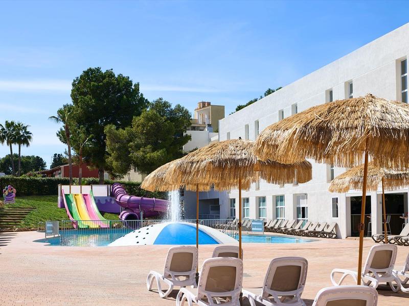 Piscina all'aperto con scivoli, ombrelloni di paglia e lettini in un hotel.