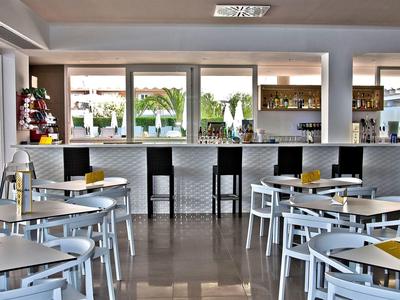 Modern hotel bar with white chairs, tables, and a black bar counter in a bright interior.