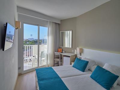 Bright hotel room with double bed, blue pillows, desk, and balcony overlooking the city.