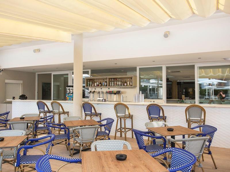 Modern outdoor hotel bar area with wooden tables and blue chairs under a bright sunshade.