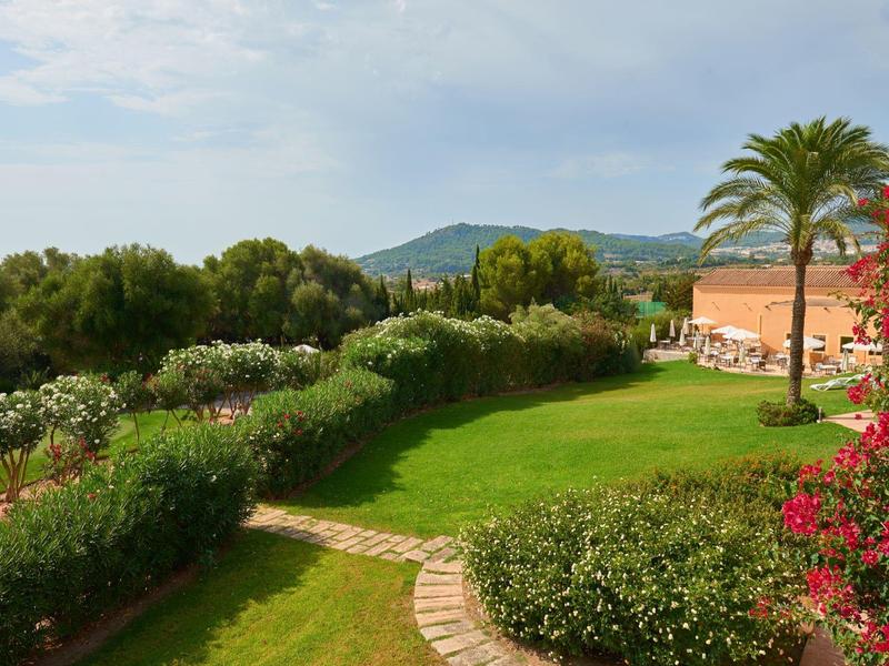 Jardin vert avec des palmiers, des fleurs colorées et vue sur les collines et la mer en arrière-plan.