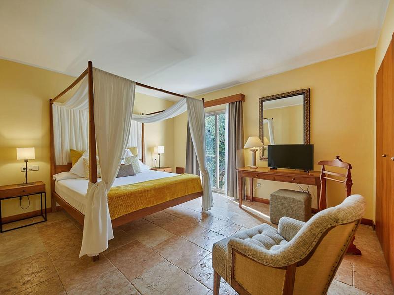 Chambre d'hôtel confortable avec lit à baldaquin, fauteuil, téléviseur et accès au balcon par portes vitrées.