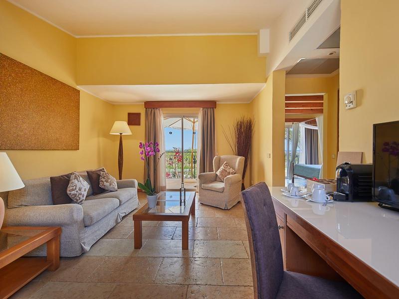 Chambre d'hôtel confortable avec canapé, fauteuil, bureau et balcon avec vue sur la verdure.