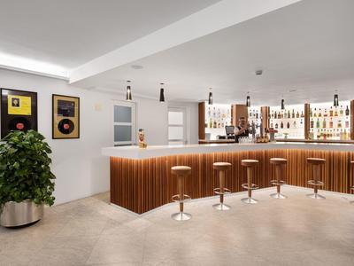 Modern hotel bar area with wooden bar, stools, and plants in bright setting.