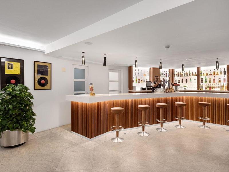 Modern hotel bar area with wooden bar, stools, and plants in bright setting.