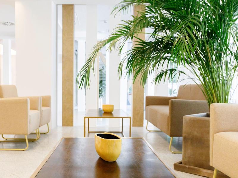 Bright, modern lobby with beige armchairs, wooden tables, and a large green plant.