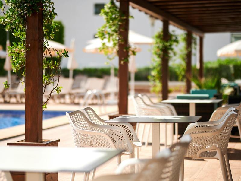 Zewnętrzny obszar z białymi wiklinowymi krzesłami i stołami przy basenie pod drewnianym pergolą.