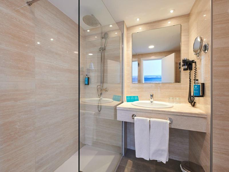 Nowoczesna łazienka z przeszklonym prysznicem, dużym lustrem, umywalką i ręcznikami w beżowych odcieniach.