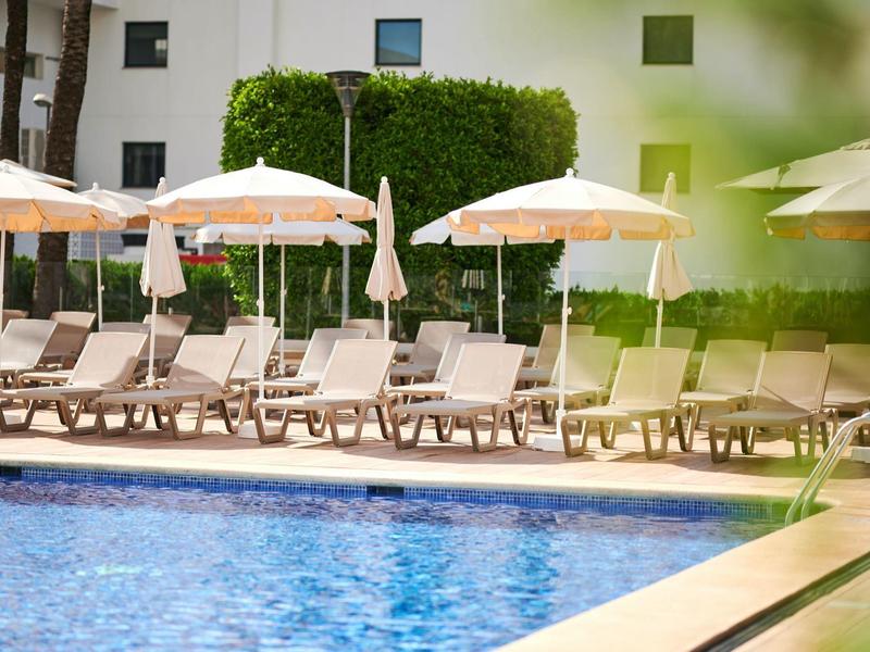 Puste leżaki z beżowymi parasolami przy niebieskim basenie i białym budynku.