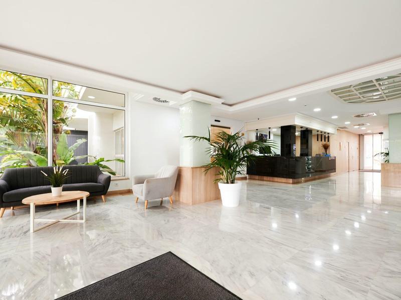 Jasny hol hotelowy z marmurową podłogą, zielonymi roślinami i nowoczesnymi siedzeniami przy dużych oknach.