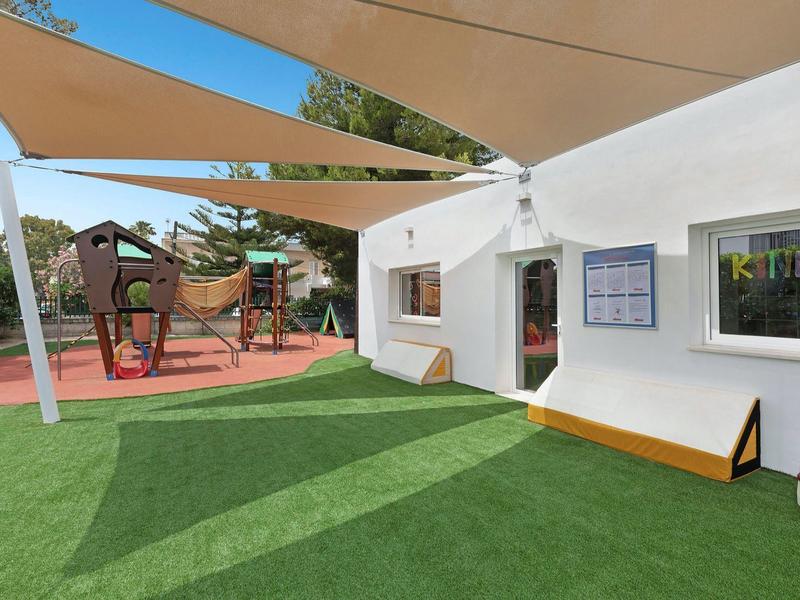 Plac zabaw z drewnianą konstrukcją do wspinaczki obok białego budynku pod beżowymi żaglami przeciwsłonecznymi