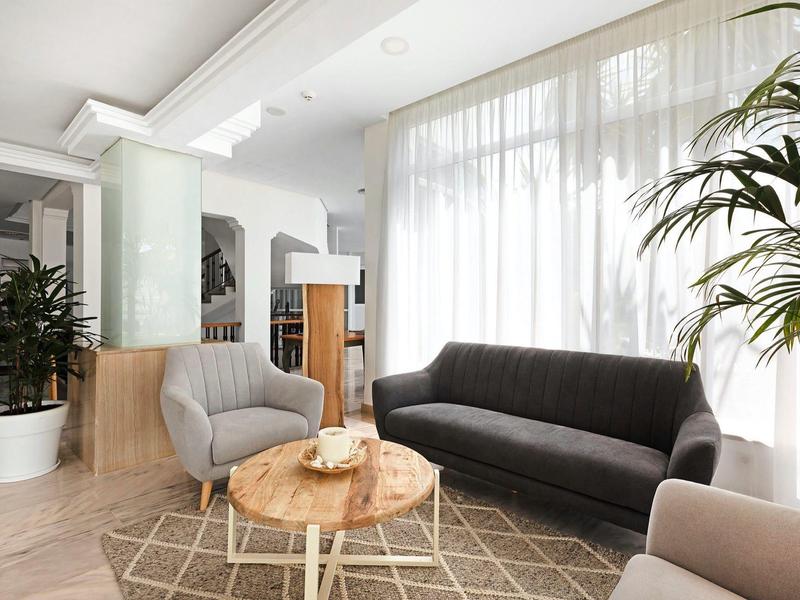 Jasny salon hotelowy z dużymi oknami, nowoczesną szarą sofą, fotelem, drewnianym stolikiem i roślinami.