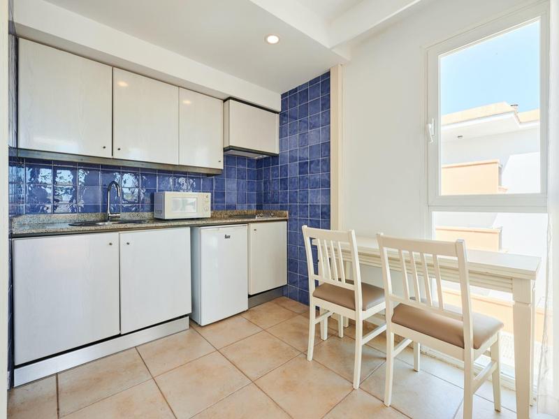 Kompaktowa kuchnia z białymi szafkami, niebieskimi płytkami, małą lodówką i stołem przy oknie