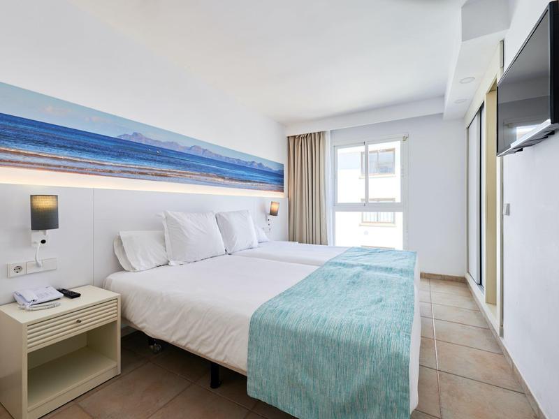 Jasny pokój hotelowy z podwójnym łóżkiem, niebieskim narzutem, szafką i telewizorem na ścianie.