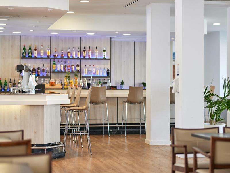 Nowoczesny bar hotelowy z drewnianą podłogą, białymi filarami i wysokimi stołkami barowymi.