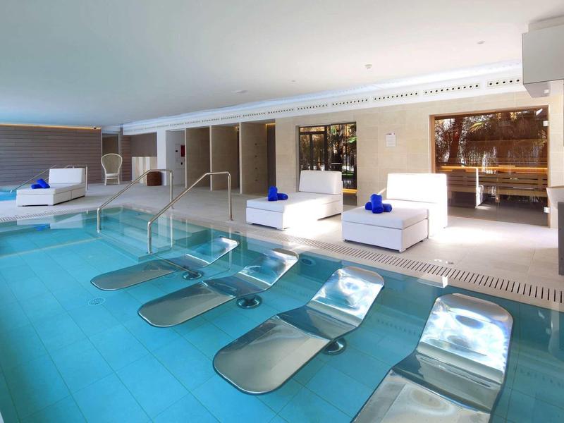 Nowoczesny basen hotelowy z wbudowanymi leżakami i białymi meblami przy basenie.