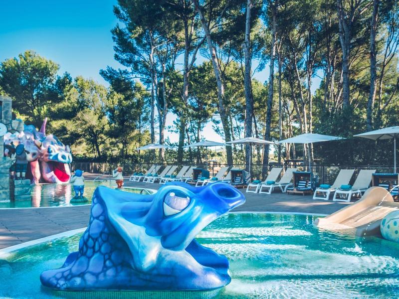 Basen dla dzieci z niebieską żabą i zjeżdżalniami obok leżaków w nasłonecznionym ogrodzie.