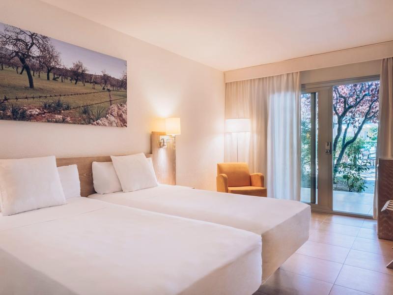 Jasny pokój hotelowy z dwoma łóżkami, fotelem i balkonem z widokiem na drzewa.