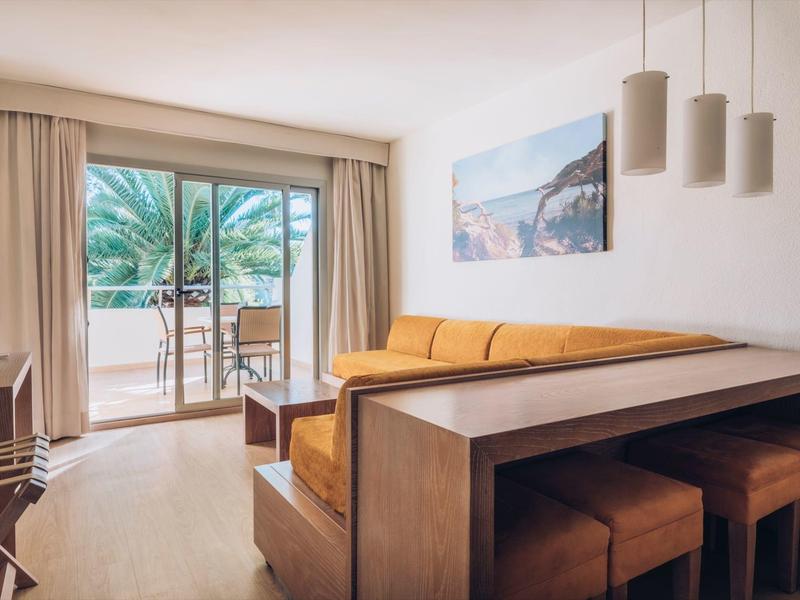 Jasny pokój hotelowy z balkonem, żółtą sofą i stołem jadalnym z krzesłami.