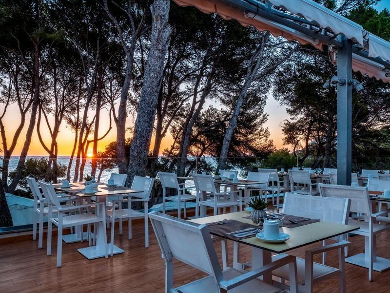 Taras restauracyjny o zachodzie słońca z widokiem na morze i drzewa.