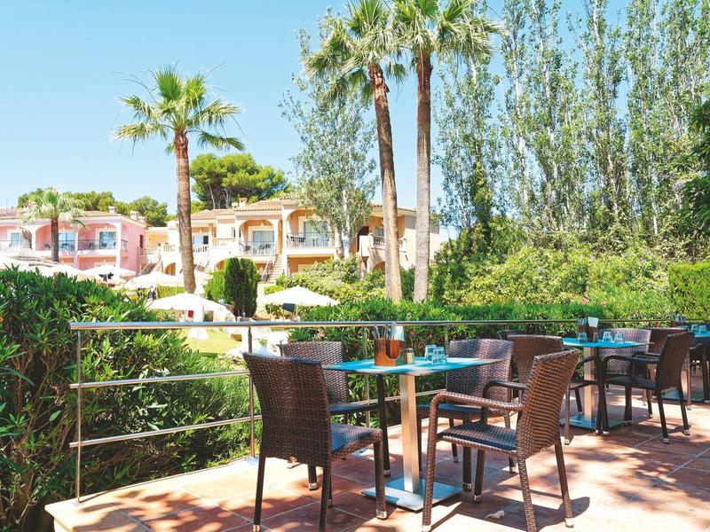 Terrazza con tavoli e sedie in un giardino con palme e edifici sullo sfondo.