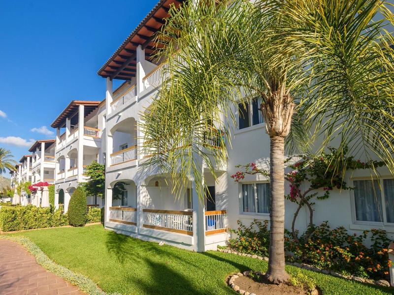 Nowoczesny kompleks hotelowy z balkonami i palmami pod jasnym niebieskim niebem.