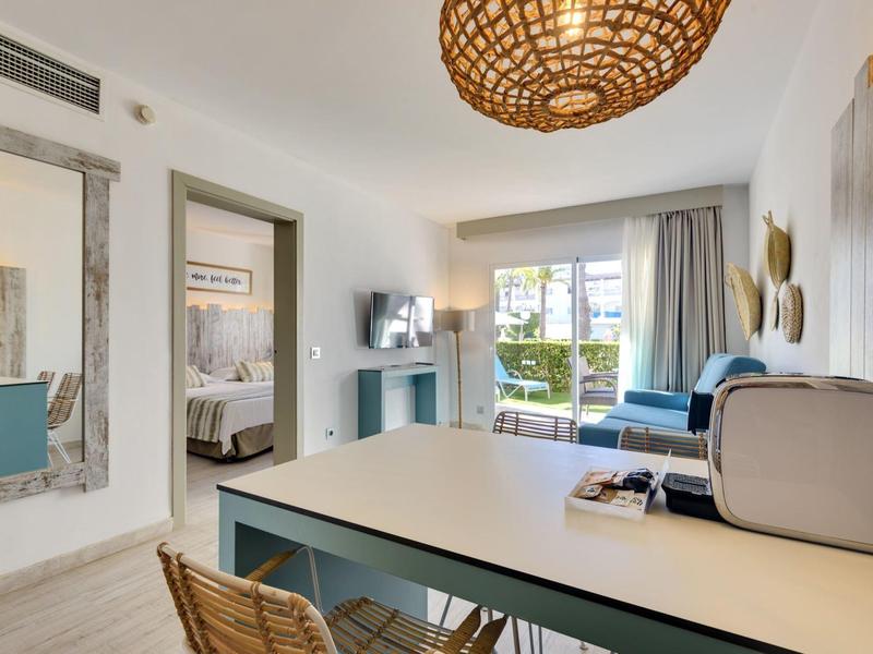 Jasny pokój hotelowy z stołem, krzesłami, sofą, telewizorem i widokiem na balkon lub taras.