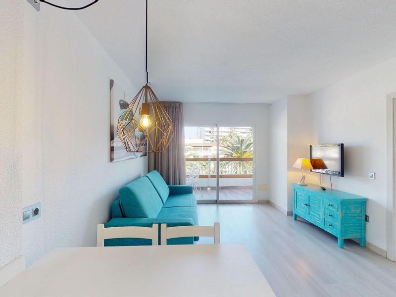Jasny, nowoczesny pokój hotelowy z turkusową sofą, komodą i przylegającą sypialnią.