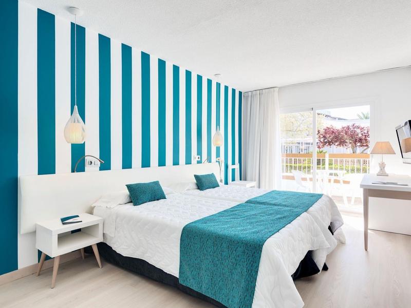 Jasny pokój hotelowy z dwoma łóżkami, niebieskimi akcentami i drzwiami balkonowymi z widokiem na ogród.