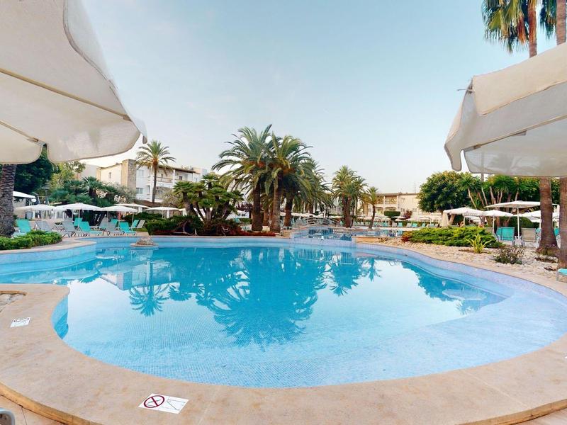 Okrągły basen hotelowy z białymi parasolami i leżakami pod niebieskim niebem.