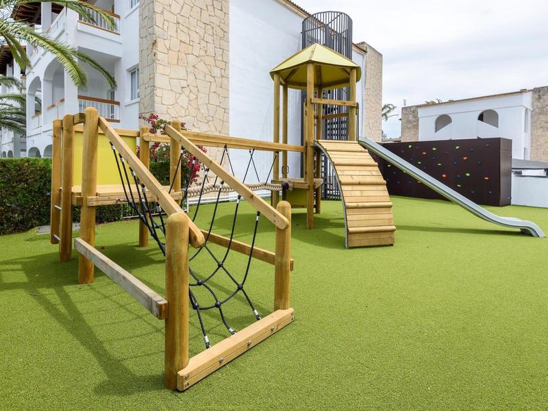Plac zabaw z drabinką i zjeżdżalnią na zielonej sztucznej trawie obok budynku.